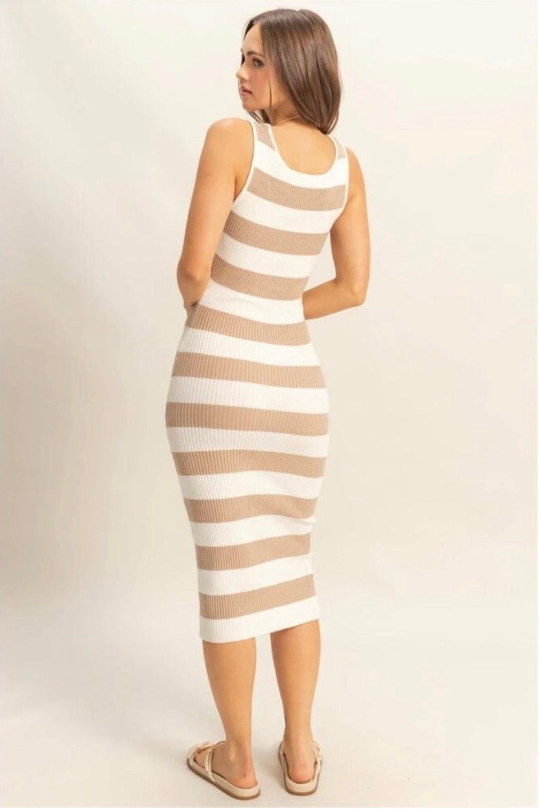 A woman modeling a sleeveless ribbed knit midi dress with horizontal beige and white stripes, shown from a rear three-quarter angle against a plain light beige studio background.